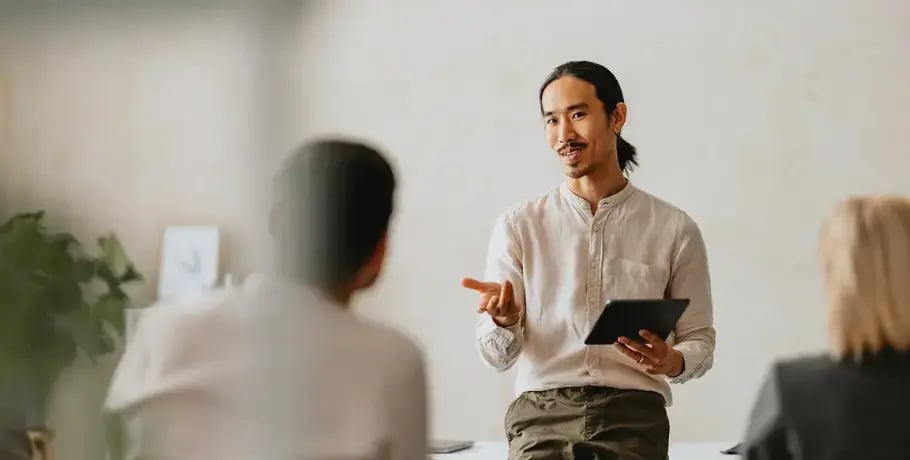 Photo of an Asian man holding a tablet and speaking to two people sat in front of him