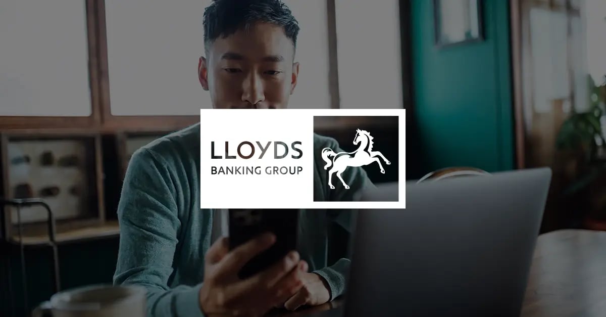 Photo of an Asian man holding a mobile phone in front of his with a white version of the Lloyds Banking Group logo superimposed in the middle foreground