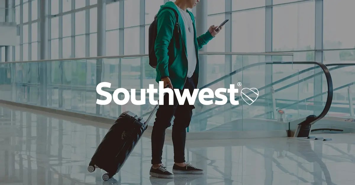 A photo of a man standing in an airport terminal with a roller suitcase and mobile phone in-hand with a white Southwest Airlines' logo in the middle