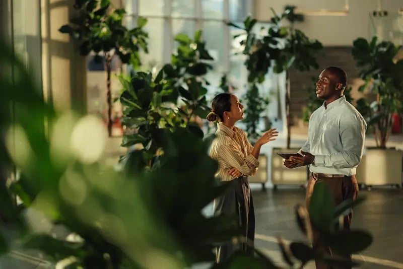 Two people having a conversation in a bright, plant-filled indoor office space. One person is holding a tablet and smiling while the other gestures as they speak.