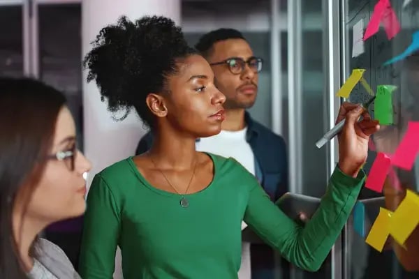 Two women and a man stood in front of a glass wall with post-it notes stuck to it and one woman writing something on one of them
