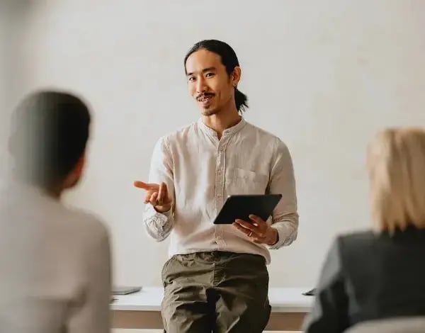 Photo of a man holding a tablet and speaking to two people sat in front of him