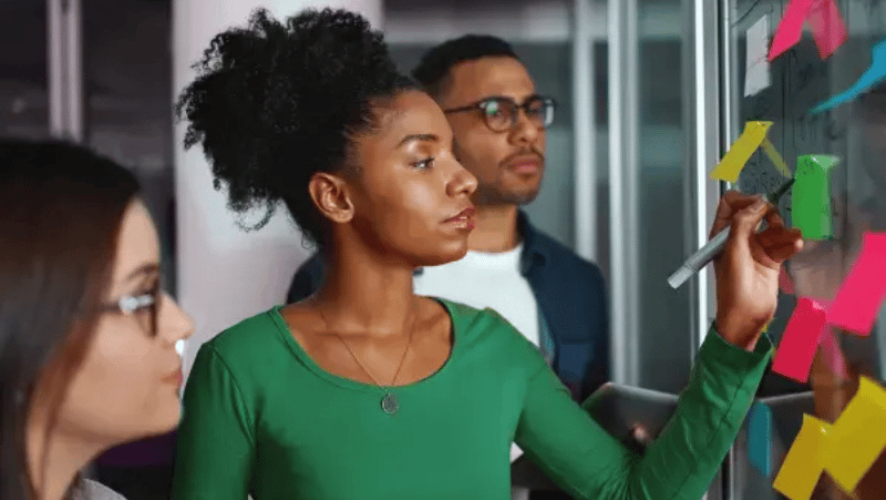 A black woman writing something on a white board with two people standing around her